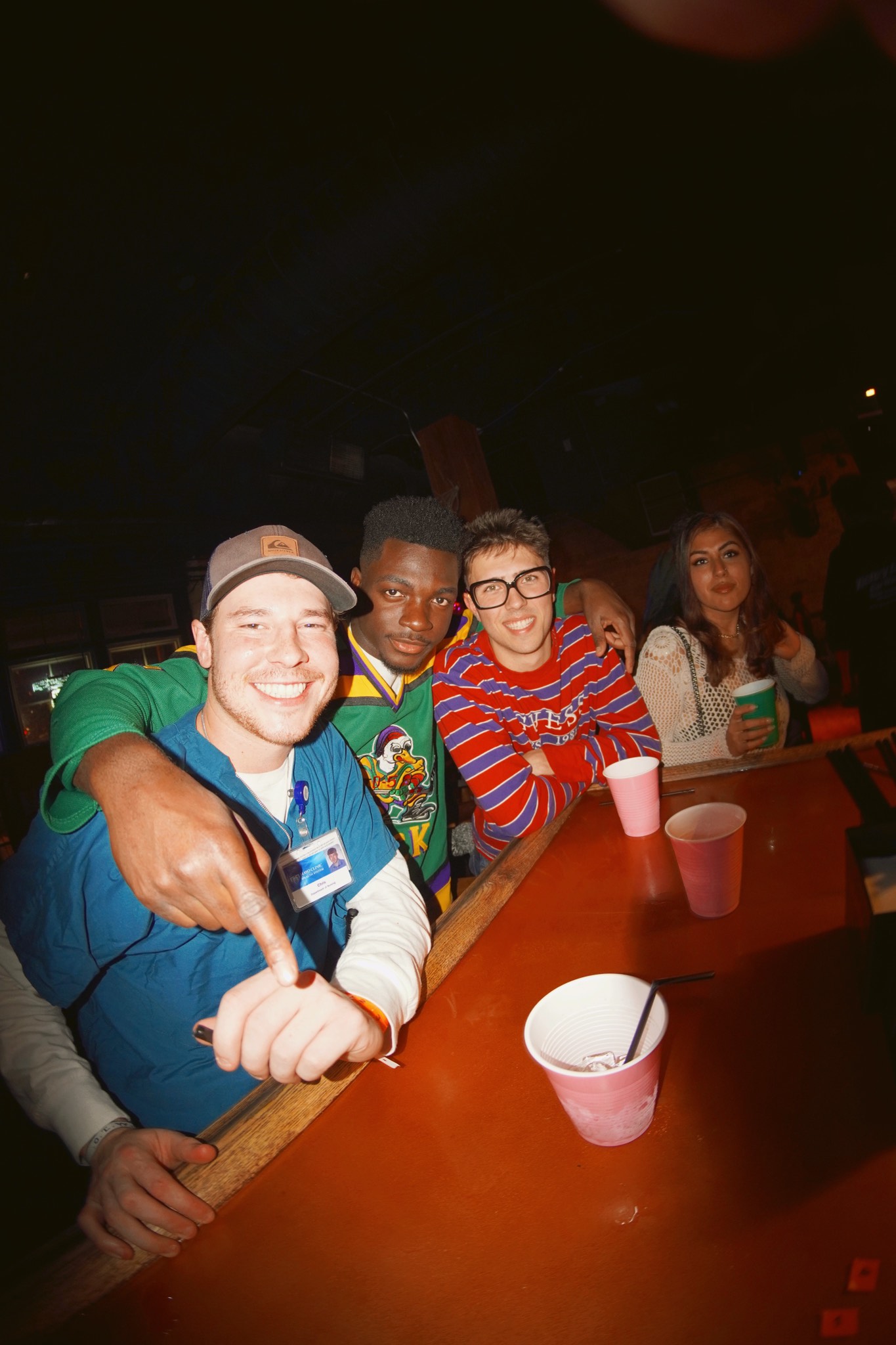 Four crawlers leaning over a bar rail in nineties costumes, one pointing straight at the camera, cups lined up on the bar in front of them