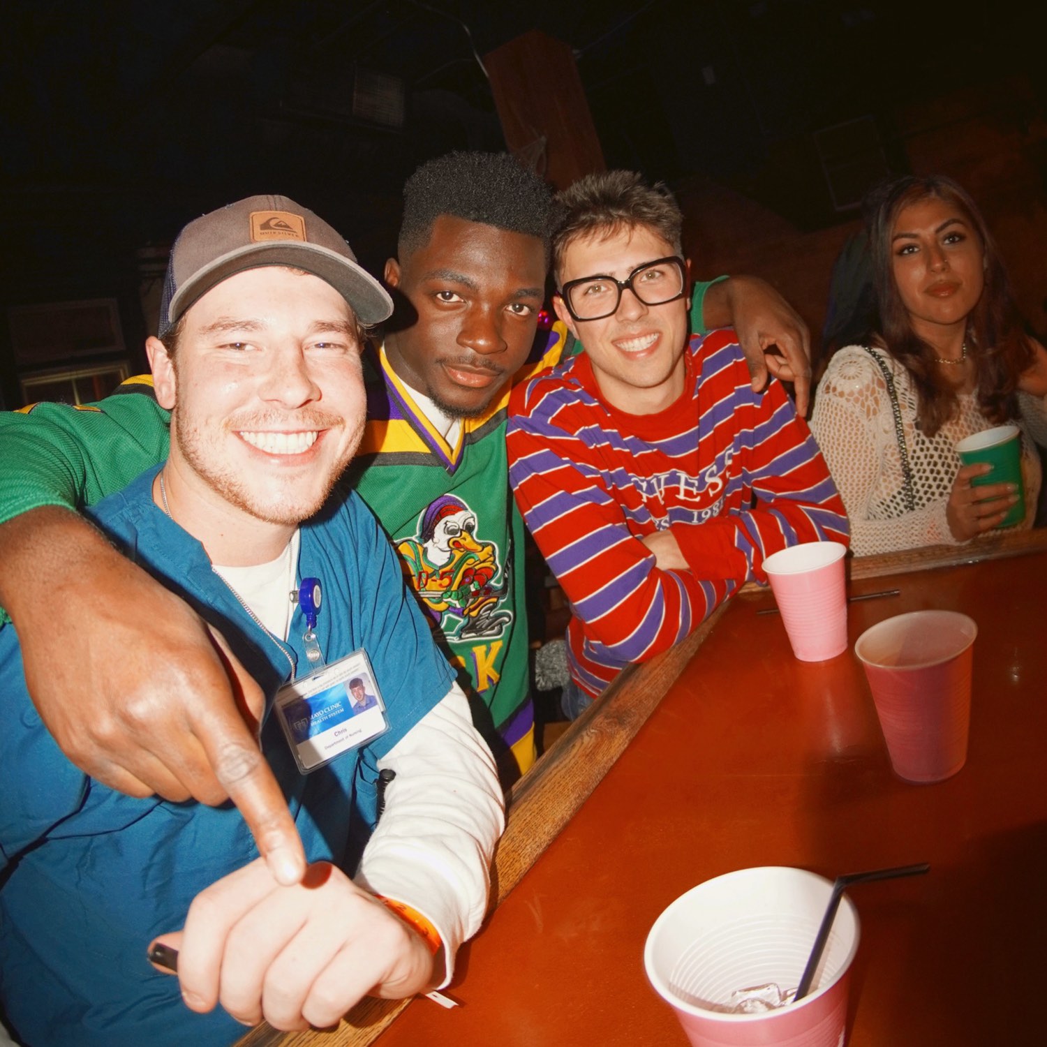 Four crawlers leaning over a bar rail in nineties costumes, one pointing straight at the camera, cups lined up on the bar in front of them