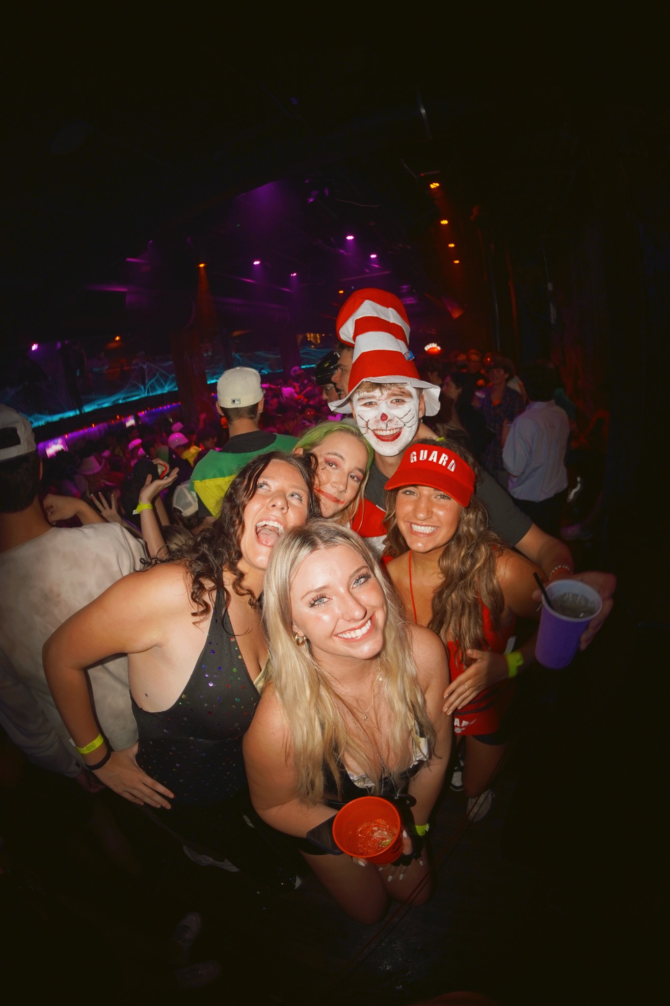 A group of crawlers packed shoulder to shoulder under purple club lighting, one in a tall striped hat and painted clown face grinning over the rest