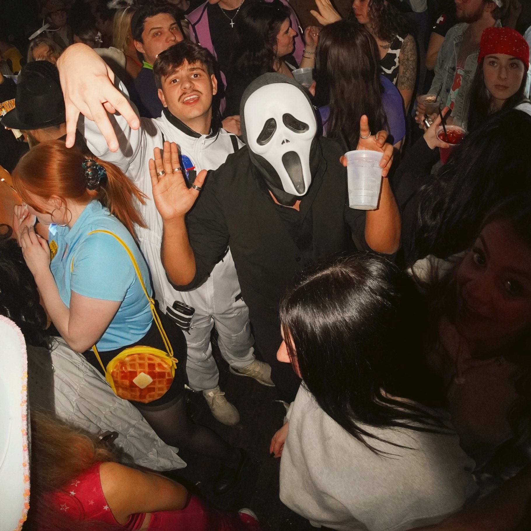 A crawler in a Ghostface mask holding a drink up in the middle of a crowded dance floor, hands raised on every side
