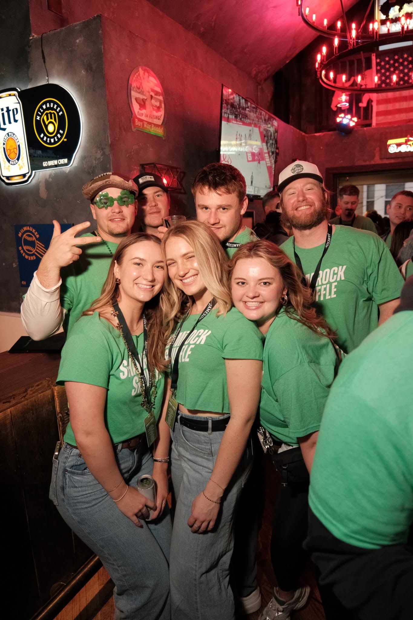 Six friends in green Shamrock Shuffle shirts crammed together for a photo at a Milwaukee bar, neon beer signs behind them