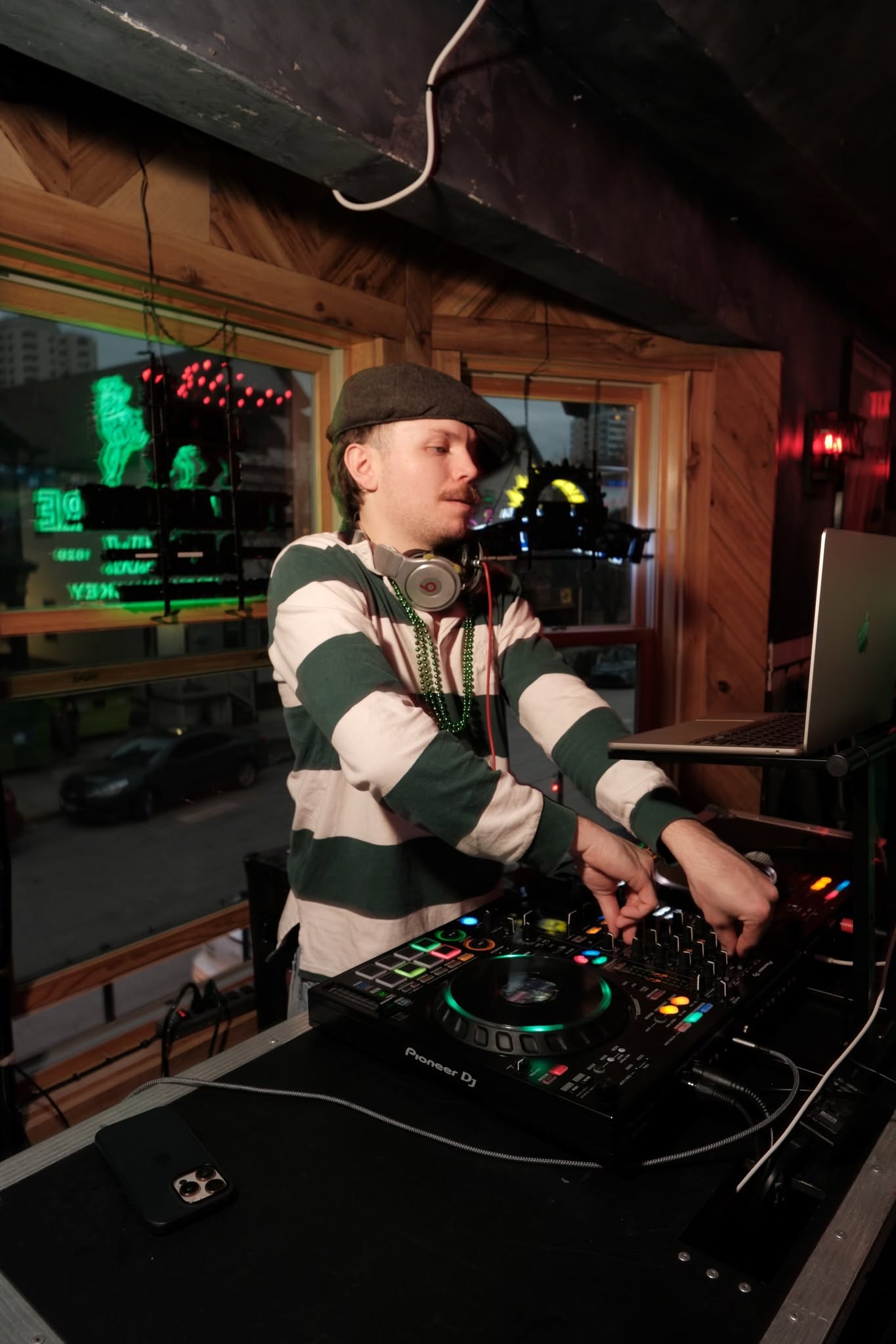 A DJ in green beads working a controller in the front window of a bar, green neon in the glass behind him